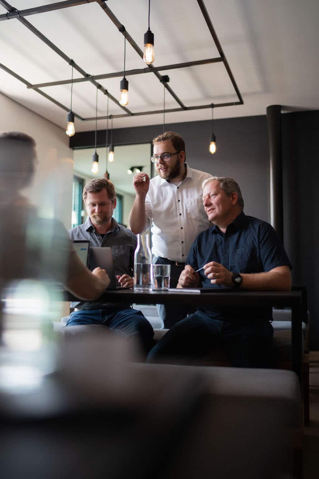 Three colleagues in a modern office lounge sit around a small table, discussing something on a laptop, with hanging lights above and water glasses on the table.