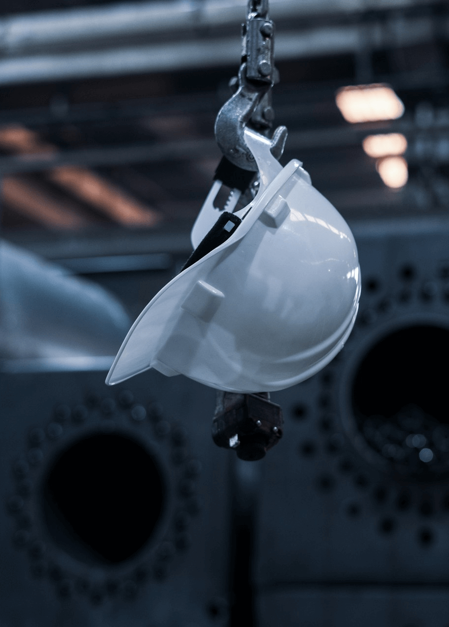 A white safety helmet hanging from a metal hook and chain inside an industrial production hall, with machinery softly blurred in the background